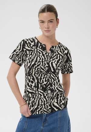 Ung kvinde med håret bundet tilbage iført sort-hvidt kortærmet top med abstrakt print og blå denim nederdel, stående med den ene hånd i lommen.