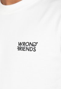 Camiseta de algodón blanca con un logo impreso en negro "WRONG FRIENDS" en el lado izquierdo del pecho. Textura suave, diseño clásico de cuello redondo.