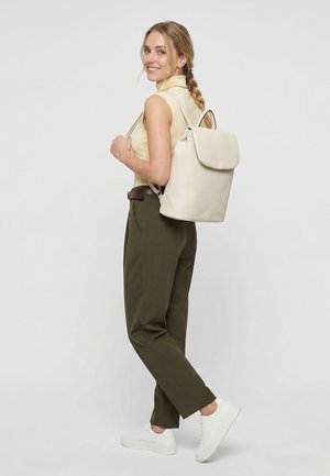 Junge Frau mit geflochtenem Haar, die ein ärmelloses Oberteil, olivgrüne Hose, weiße Sneakers trägt und einen hellbeigen Rucksack trägt, lächelt zurück.