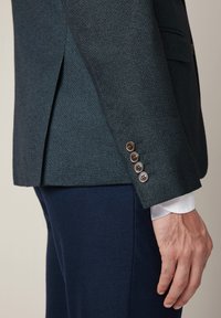 Zbliżenie na osobę w teksturowanej ciemnozielonej marynarce z czterema brązowymi guzikami przy mankietach i granatowymi spodniami, z ręką swobodnie opartą przy boku.