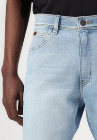 Jeans de ganga de cor azul clara com uma cintura alta, com fecho de botão, costura discreta e uma etiqueta de couro no bolso de trás.