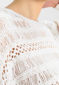 Detailní záběr na osobu v bílém pleteném topu s třásněmi a otvorovými vzory, ktrerý odhaluje krk a horní část ramen.