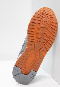 Schoenzool met oranje en grijze rubberen profiel, "New Balance" en "XAR 1000" merk zichtbaar, schuin tegen een witte hoekoppervlakte.