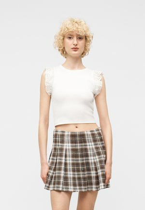 Jeune femme aux cheveux blonds courts et bouclés portant un haut blanc sans manches à volants et une jupe plissée à carreaux marron.