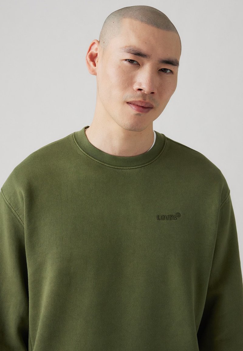 Sweatshirt vert olive avec un col rond, des poignets côtelés et un petit logo en relief sur la poitrine gauche, fabriqué en tissu en coton texturé.