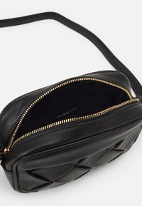 Sort quiltet læder crossbody taske med gylden lynlås delvist åben, der viser stofinteriør og justerbar rem på hvid baggrund.