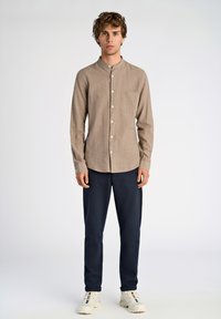 Camisa de lino color marrón claro con botones, cuello y un bolsillo en el pecho, combinada con pantalones azul marino y zapatillas blancas.