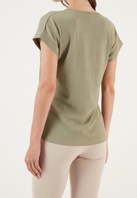 T-shirt verde oliva chiaro con finitura texturizzata, maniche corte arrotolate e vestibilità rilassata. Indossato con leggings beige per un contrasto.