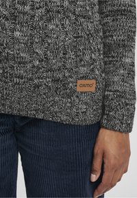 Pull en maille gris-noir avec un motif de câble texturé et une étiquette en cuir beige affichant "OXMO". Assorti à un pantalon en velours côtelé bleu marine.