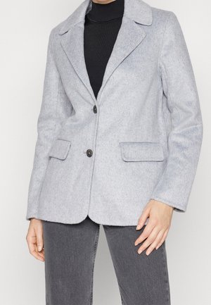 Frau trägt hellgrauen Wollblazer mit zwei Knöpfen, Pattentaschen, über schwarzem Rollkragenpullover und dunkelgrauen Jeans, steht mit einer Hand nach unten.