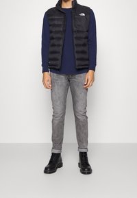 Černá polstrovaná vesta s vysokým límcem přes tmavě modrou košili s dlouhým rukávem, spárovaná se šedými slim fit džíny a černými šněrovacími botami.