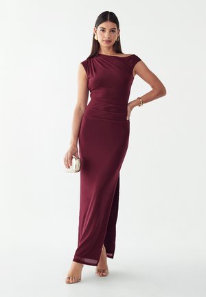 Vrouw draagt een mouwloze bordeaux avondjurk en houdt een kleine zilveren clutch vast, met gouden oorbellen, armband en open sandalen.