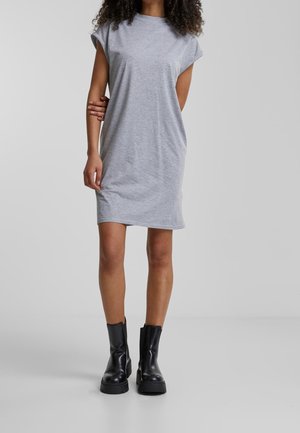 Femme portant une robe grise à manches courtes et des bottines noires épaisses, debout devant un fond uni.