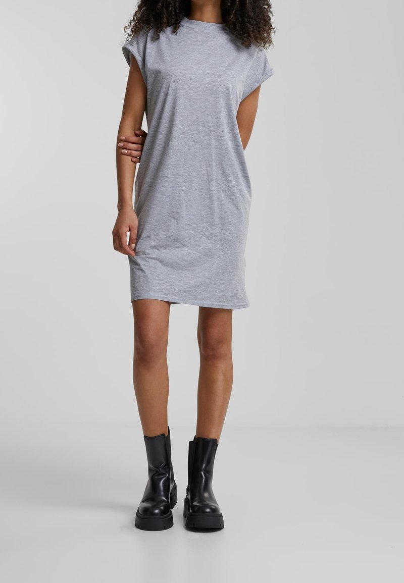 Femme portant une robe grise à manches courtes et des bottines noires épaisses, debout devant un fond uni.