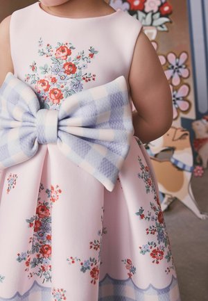 Child wearing a pink floral dress with a large blue and white checkered bow on the back, standing indoors near painted wall art.