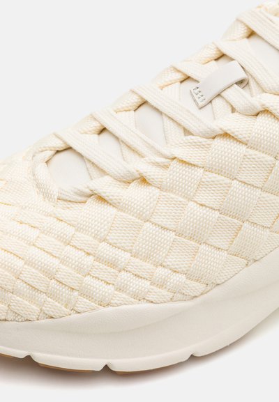 Sneaker di colore crema intrecciato con materiale testurizzato, lacci piatti e suola bianca leggera con finitura liscia.