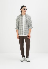 Conjunto masculino que incluye una camisa de cuadros gris, una camiseta blanca, pantalones marrones ajustados y unas zapatillas blancas/plateadas con detalles en negro.
