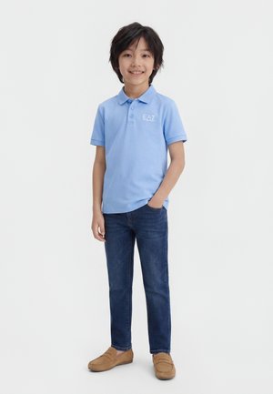 Fant z roko v žepu, oblečen v svetlo modro polo majico, temne kavbojke in rjave usnjene čevlje, se smeji v kamero.