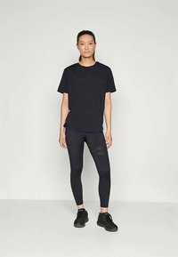 Egy nő áll, arccal előrefelé nézve, fekete, oversized pólót, fekete csíkos leggingst és fekete sportcipőt visel egy sima, világos háttér előtt.