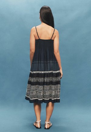 Femme aux longs cheveux foncés portant une robe sans manches à motif noir et blanc et des sandales blanches, debout devant un fond bleu, de dos.