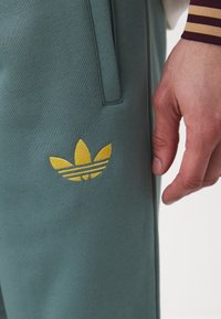 Green joggers made of ribbed fabric featuring a yellow embroidered Adidas logo with three stripes near the pocket. Close-up detail shown.