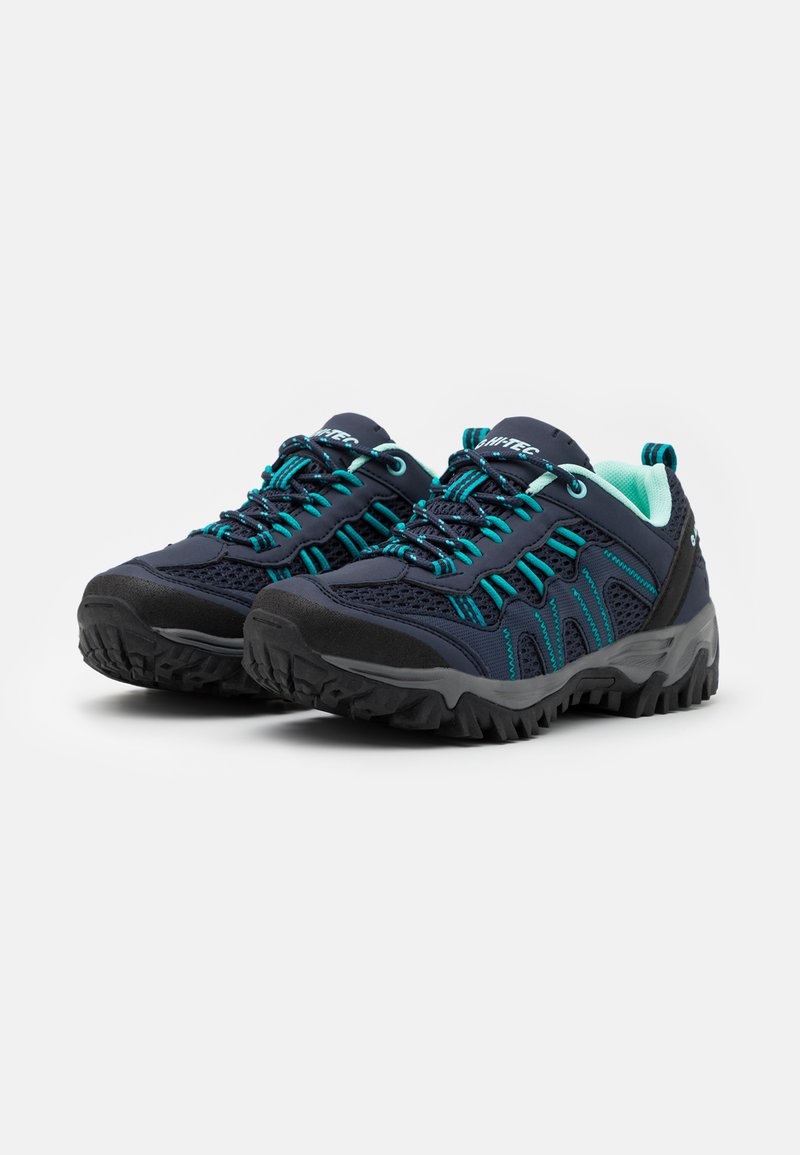 Zapatos de senderismo azul oscuro con inserciones de malla, acentos en azul verdoso, suelas de goma negra y agarres texturizados. Presentan un sistema de cordones prominente y un lazo en la parte trasera.