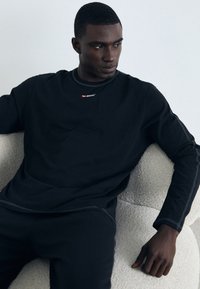Black long-sleeve top with rounded neckline, featuring a small red "DIESEL" logo. Smooth fabric with contrast stitching, worn with matching black pants.