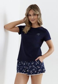 Granatowa koszulka na krótki rękaw z logiem, w połączeniu z szortami w kwiatowy wzór z koronkowym wykończeniem. Wygodny, casualowy zestaw odzieży do domu.