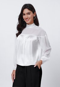 White satin blouse with a high neckline, long sleeves, and gathered detailing. Smooth texture, designed with a relaxed fit.
