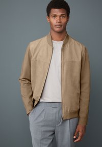Beige Bomberjacke mit glatter Textur und gerippten Bündchen, getragen über einem hellgrauen Hemd. Das Modell steht vor einem einfarbigen grauen Hintergrund.
