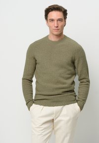 Maglione lavorato a maglia verde oliva con scollo rotondo, superficie testurizzata e polsini a costine. Abbinato a pantaloni di colore crema.