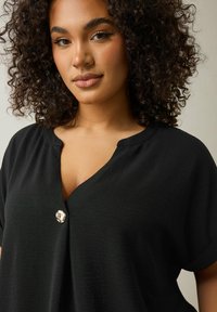 Top nero a maniche corte con scollatura a V e un bottone decorativo argento. Il tessuto ha un aspetto liscio e leggermente testurizzato.