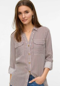 Chemise à boutons avec des rayures verticales bordeaux et blanches, deux poches poitrine, manches retroussées et boutons marron. Texture en tissu doux et tissé.