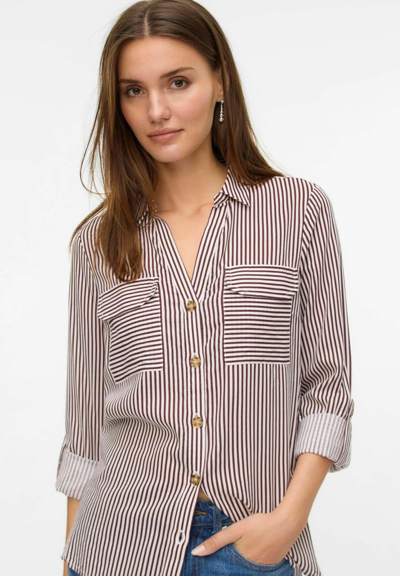 Chemise à boutons avec des rayures verticales bordeaux et blanches, deux poches poitrine, manches retroussées et boutons marron. Texture en tissu doux et tissé.