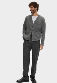 Grauer Wollcardigan mit V-Ausschnitt, vorderem Knopfverschluss und zwei Taschen, getragen über einem weißen Hemd und kombiniert mit grauen Hosen und schwarzen Schuhen.