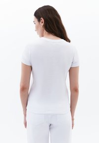 T-shirt bianco in cotone con maniche corte, scollatura tonda e texture liscia, visto da dietro, abbinato a pantaloni bianchi.