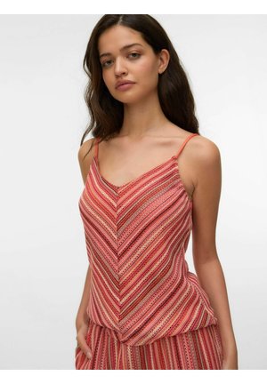 Kvinde med langt mørkt hår iført en ærmeløs, pink og rødstribet V-hals top og matchende bukser, stående med den ene hånd i lommen.