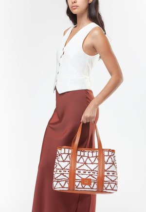 Donna con gilet bianco senza maniche e gonna marrone che tiene una borsa tote bianca e marrone con tracolle in pelle.