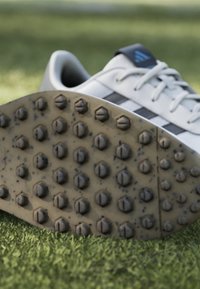 Vit atletisk sko med svarta och blå detaljer. Texturerad gummisula med framträdande hexagonala och cirkulära nubb för bra grepp.