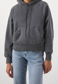 Felpa grigia con cappuccio, tasca frontale, polsini e orlo a costine. Indossata con jeans azzurri chiari con effetto usurato. Tessuto morbido, design casual.