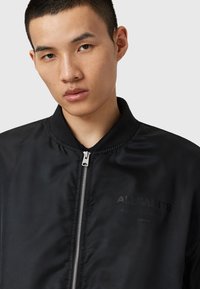 Jeune homme portant une veste zippée noire avec le logo "ALLSAINTS" sur la poitrine, sur fond gris clair uni.