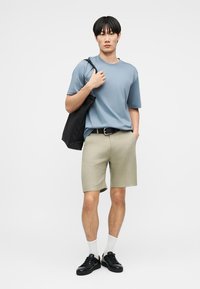 Νεαρός άνδρας με μπλε κοντομάνικο πουκάμισο, μπεζ σορτς, λευκές κάλτσες, μαύρα αθλητικά παπούτσια, που κρατά μαύρη τσάντα ώμου σε λευκό φόντο.