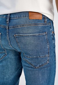 Niebieskie jeansy denimowe z beżową skórzaną naszywką z napisem "LINDBERG" na pasku, kontrastującym przeszyciem i standardowymi tylnymi kieszeniami.