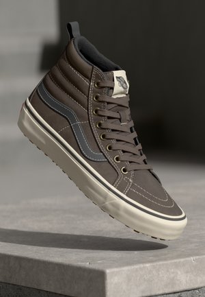 Baskets montantes Vans marron avec bande grise sur le côté et semelle blanc cassé flottant au-dessus d'une surface en béton dans un éclairage neutre.