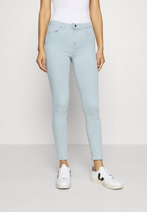 Kobieta ubrana w jasnoniebieskie obcisłe jeansy, białe trampki z czarnym logo oraz białą, wciągniętą bezrękawnik, stojąca na tle jednolitego tła.