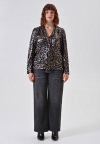 Donna con capelli ondulati fino alle spalle, che indossa una blusa lucida con stampa leopardata, jeans neri a gamba larga e stivaletti neri, in piedi su uno sfondo neutro.