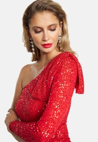 Vestito rosso decorato con paillettes, con una spalla scoperta e design texturizzato. Accentuato da un dettaglio a fiocco sulla manica.