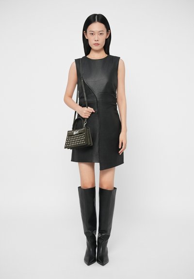 Mulher vestindo um vestido de couro preto sem mangas, botas pretas até o joelho e segurando uma mala preta com tachas, em um fundo branco.