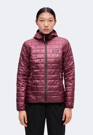 Jeune femme portant une veste matelassée à capuche bordeaux avec fermeture éclair devant et pantalon noir, debout devant un fond gris.