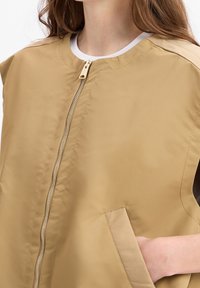 Beige Reißverschluss-Weste mit rundem Ausschnitt, ausgestattet mit Seitentaschen und glattem, leichtem Stoff. Mit einem kontrastierenden weißen Kragen.